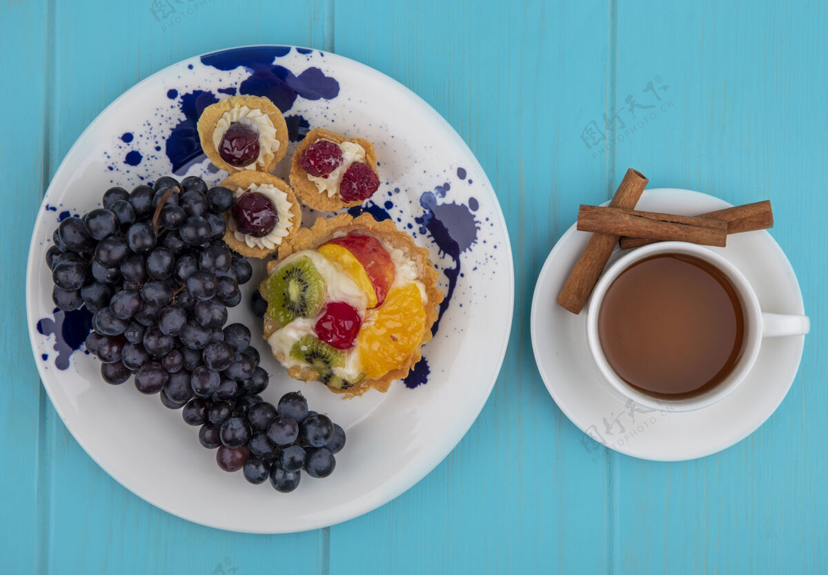 盘子盘子上的黑葡萄顶视图 蓝色木制背景上有一杯茶和肉桂棒杯子茶肉桂