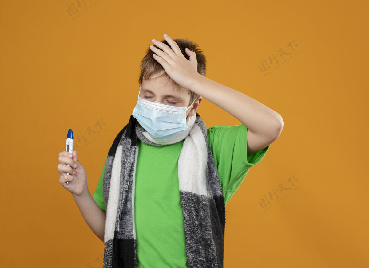 橙色生病的小男孩穿着绿色t恤 脖子上围着暖和的围巾 戴着面罩 手里拿着温度计 站在橙色的墙上 看起来很困惑看站着男孩