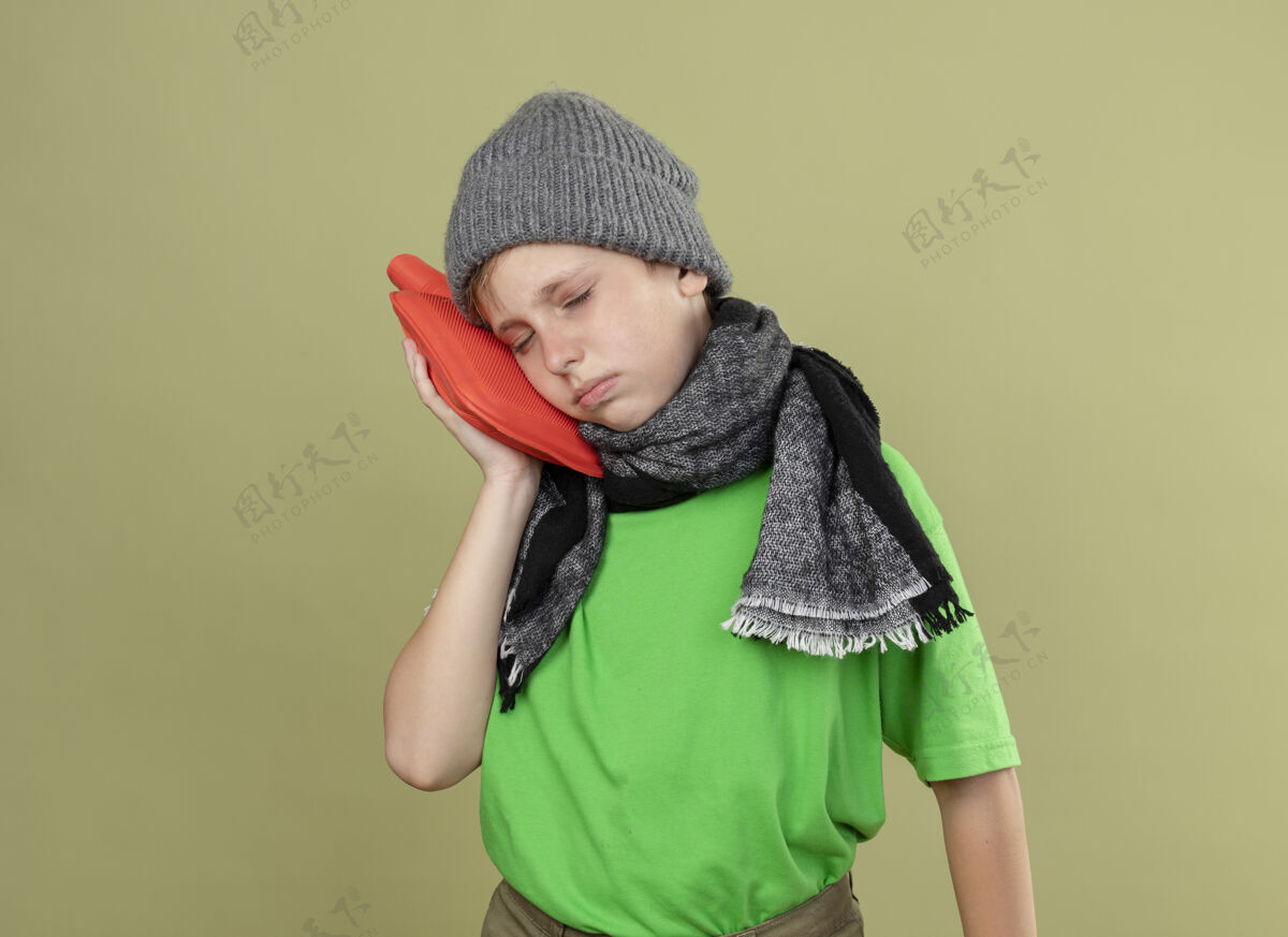 持有生病的小男孩穿着绿色t恤 戴着暖和的围巾和帽子 感觉不舒服 拿着水瓶取暖 把头靠在上面 站在轻墙上保持头围巾