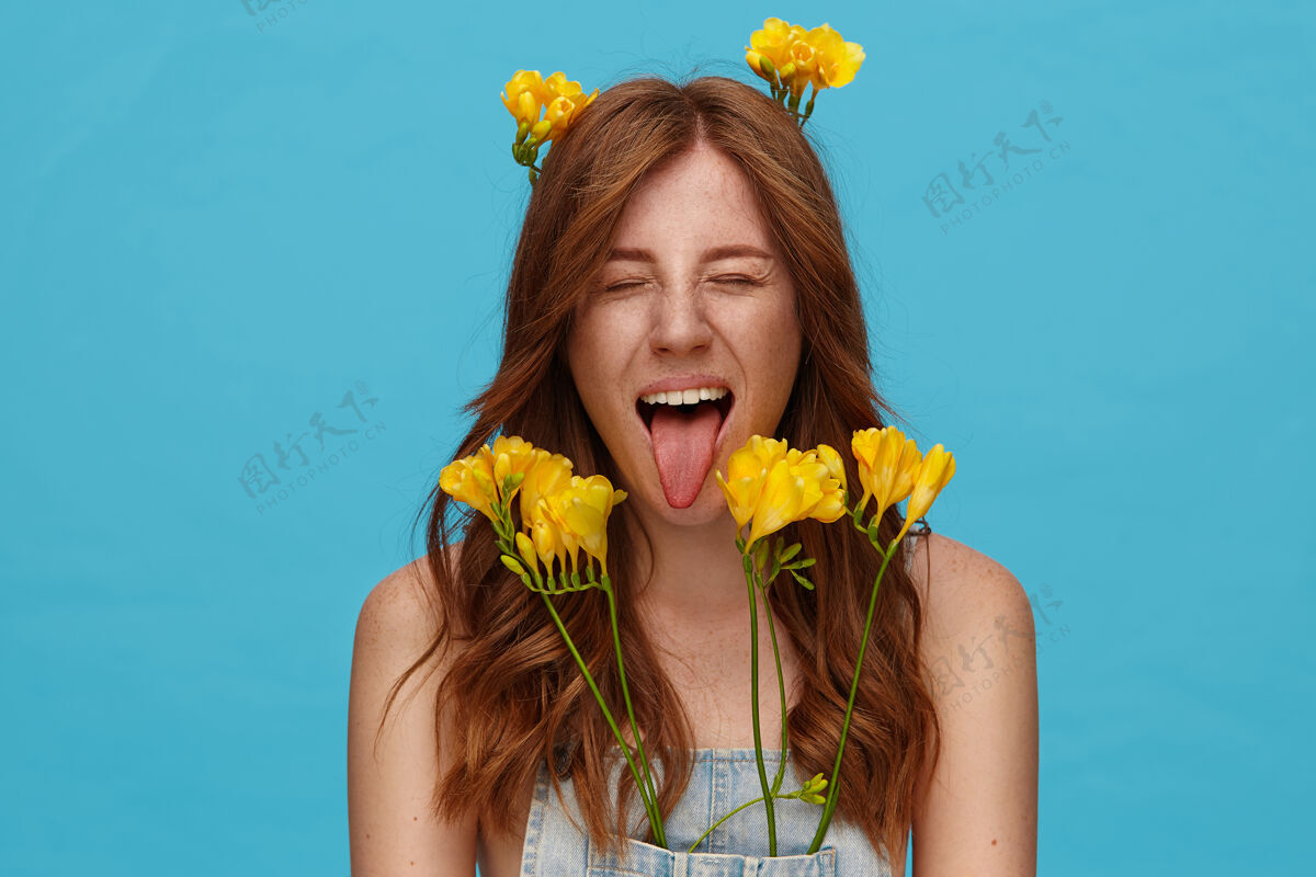 鬼脸一张室内照片 一个年轻漂亮的狐狸精女人 留着卷发 闭着眼睛 伸出舌头 在蓝色背景上摆着黄色的花自然红发年轻