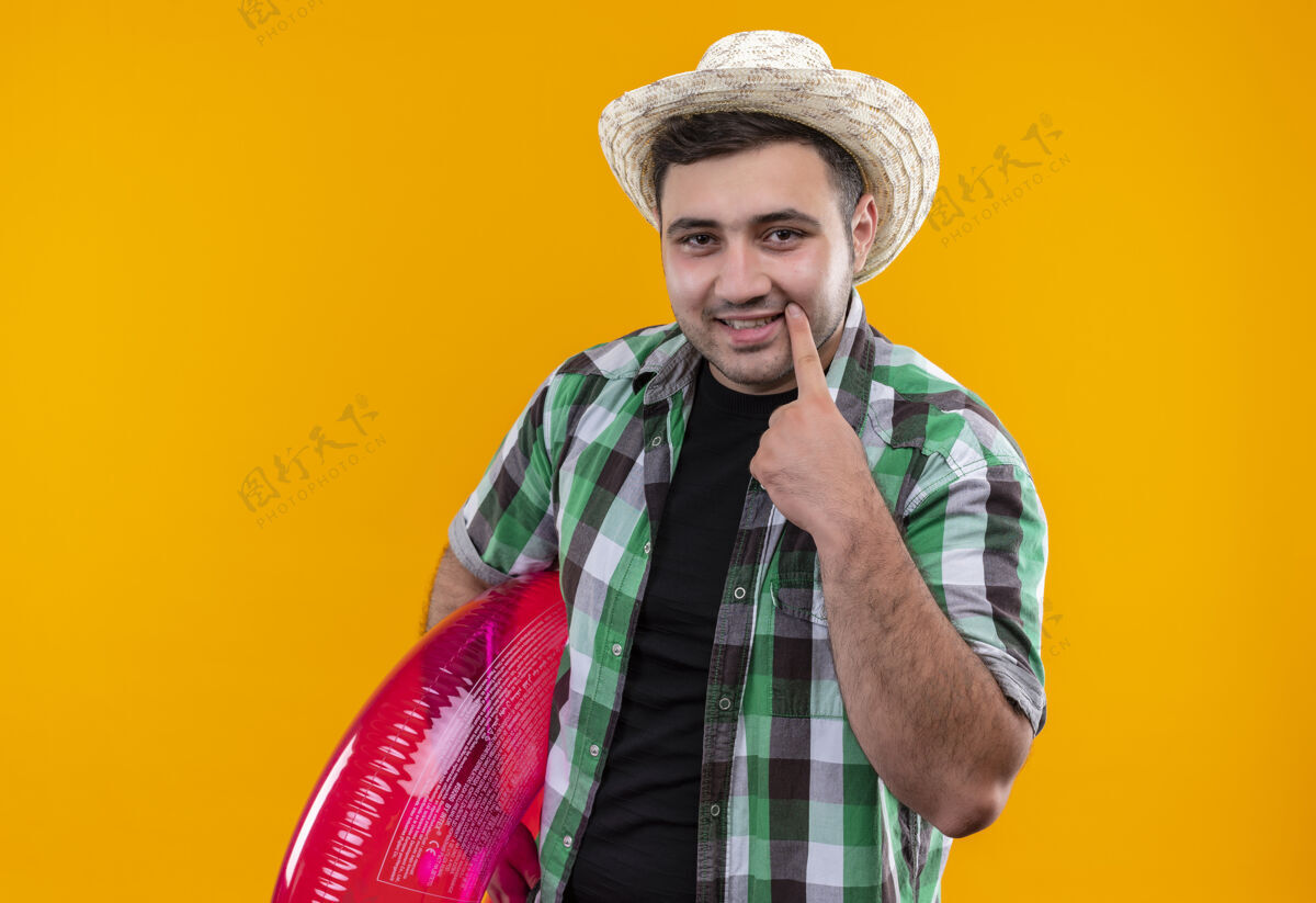 衬衫穿着格子衬衫 戴着夏帽的年轻旅行家 手拿充气戒指 手指指着他站在橘色墙上的笑容检查男人帽子