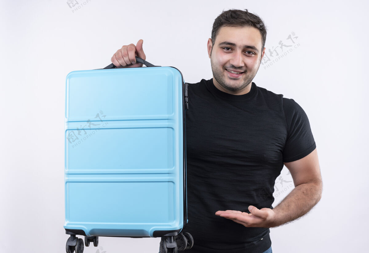 自信身穿黑色t恤的年轻旅行家 手拿手提箱 站在白色的墙边 自信地微笑着展示旅行微笑假期