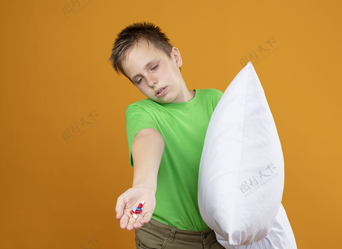感觉生病的小男孩 穿着绿色t恤 感觉不舒服 抱着枕头 手里拿着药片 站在橘色的墙上 看起来很困惑 不开心 生病生病橘子不开心