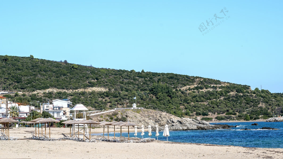岩石爱琴海海岸左侧有建筑物 岩石 带日光浴床的雨伞 灌木丛和树木 蓝色的海水和希腊萨蒂的小山海海岸悬崖