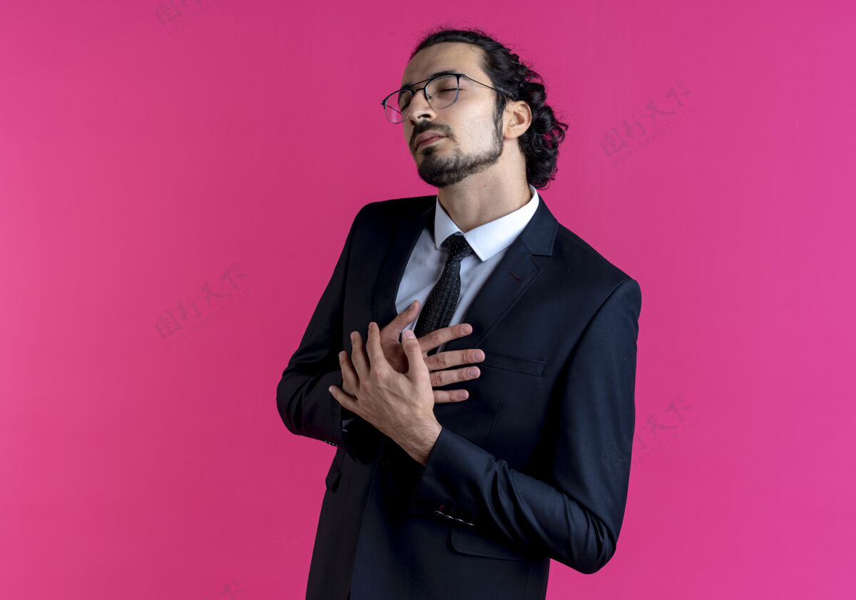 看一位身穿黑色西装 戴着眼镜的商人双手交叉放在胸前 站在粉色的墙上 一边看一边 心里充满感激姿势生意人生意
