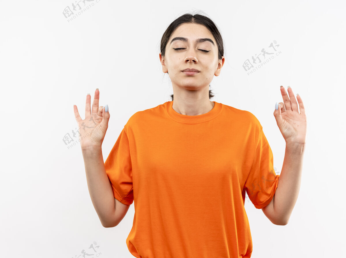 佩戴穿着橙色t恤的年轻女孩闭着眼睛放松 手指站在白墙上做冥想手势制造闭合女孩