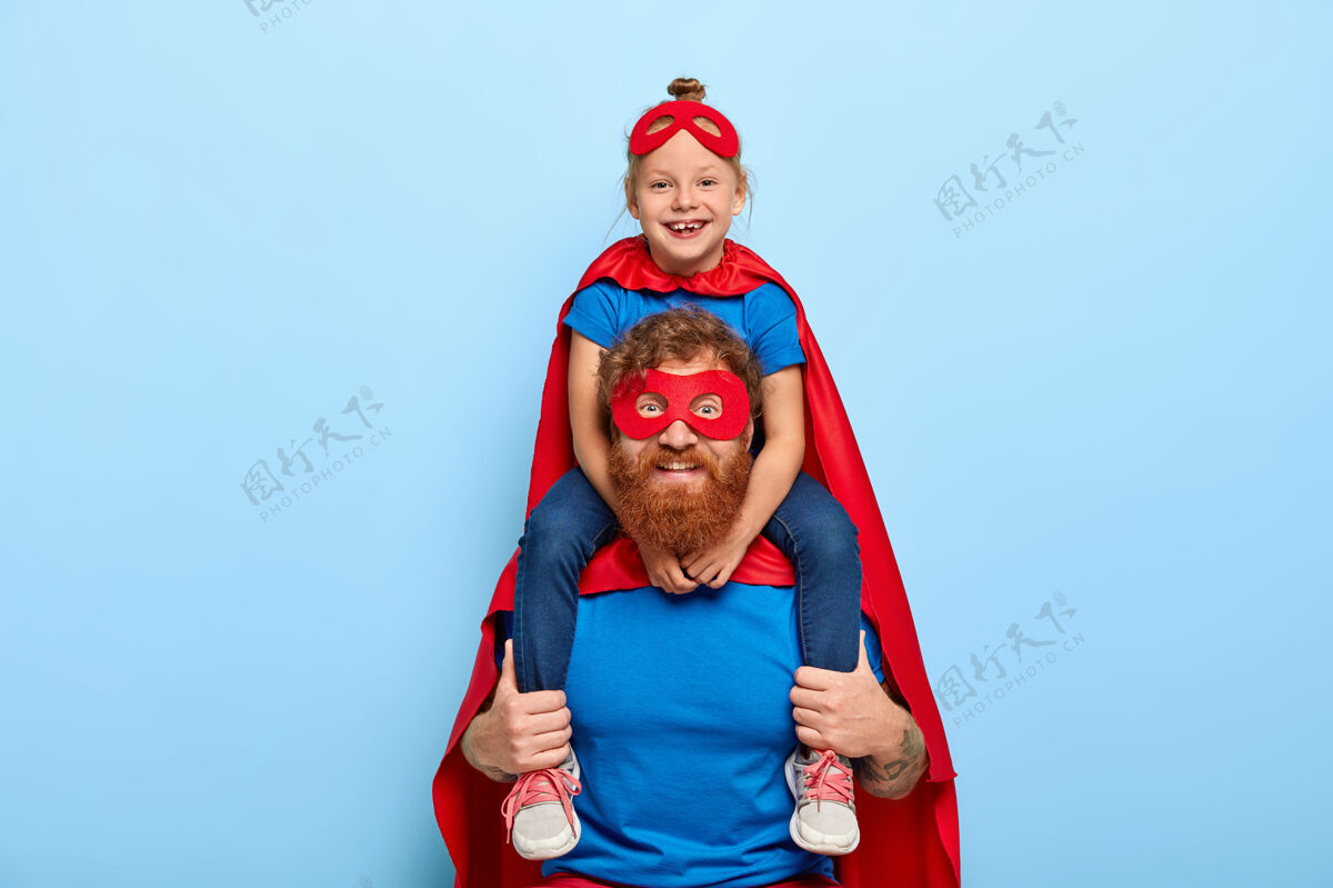 红发快乐的女孩儿坐在父亲的肩膀上 感觉英勇坚强 戴着红色的面具玩耍胡须女儿