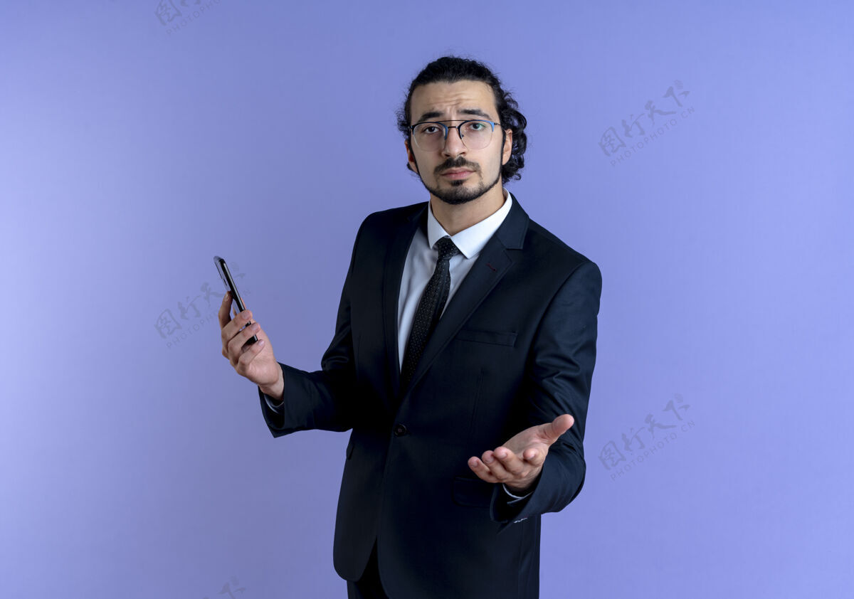 站着一位身穿黑色西装 戴着眼镜的商人举着智能手机 伸出手臂望着前方 站在蓝色的墙上问道手机商务人员看着