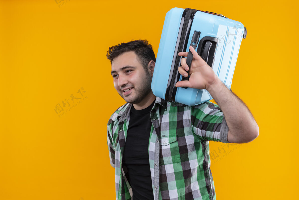 姿势身穿格子衬衫的年轻旅人肩上扛着手提箱 站在橘色的墙上 脸上带着幸福的笑容假期旅行假期