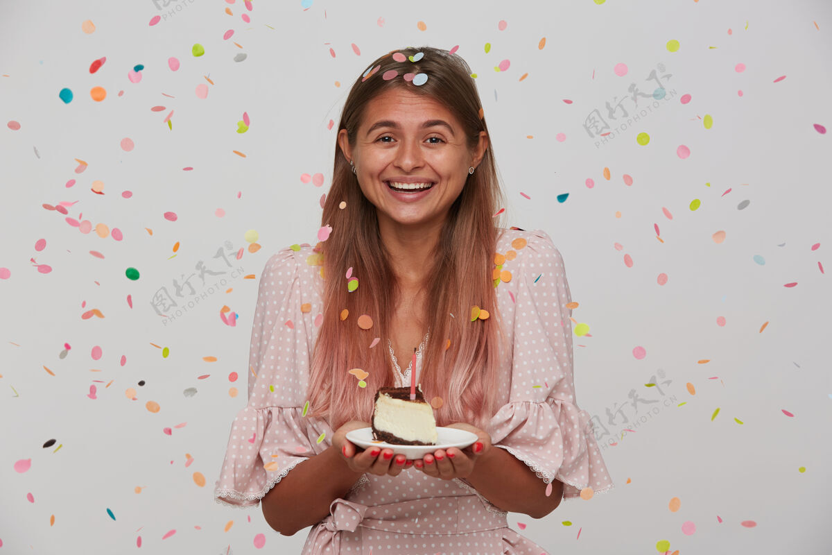 五彩纸屑快乐的年轻漂亮的长发女性在庆祝生日的时候展现出她愉快的情绪 举起蛋糕积极地微笑 在白色的墙上摆姿势节日表情缪