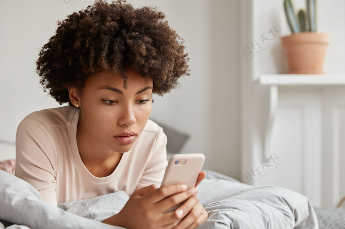 手机一位留着非洲发型的黑人年轻女士在社交网络上查看最新信息 在手机上分享多媒体文件 穿着休闲睡衣 在床上休息 在线支付休息更新黑色