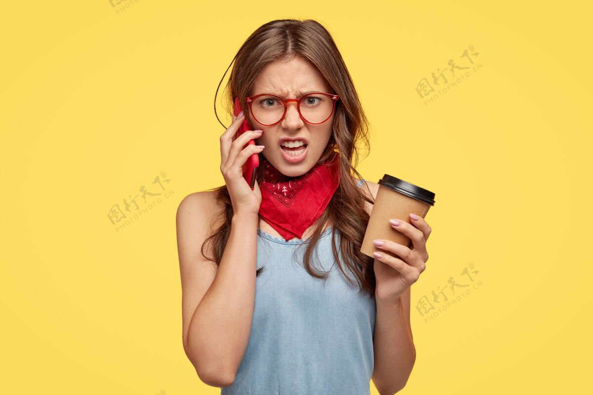 恼怒愤怒的年轻女子在和朋友通电话时感到紧张 听到废话 不同意某事 不喜欢地笑 喝咖啡 隔着黄墙孤立交谈皱眉惊奇