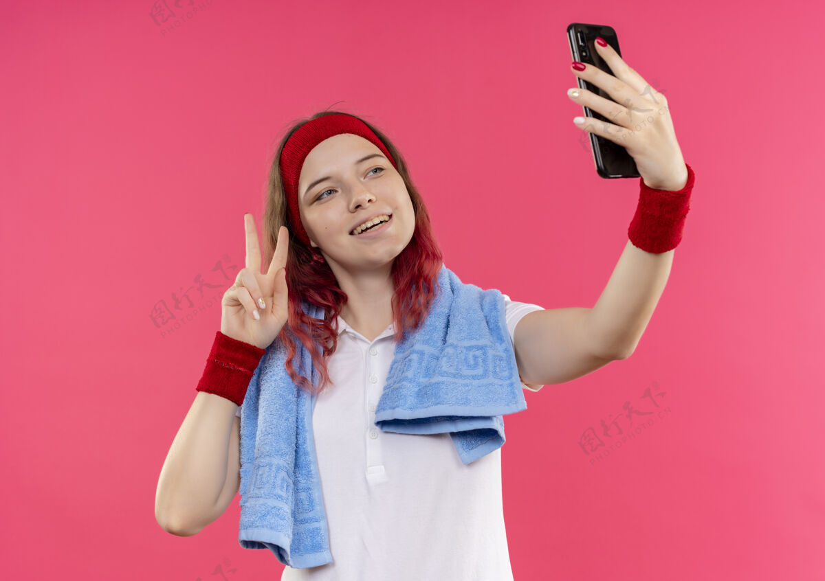 胜利戴着头巾肩上拿着毛巾的年轻运动女性自拍 在粉色墙上对着智能手机的镜头展示胜利标志她自己运动装人