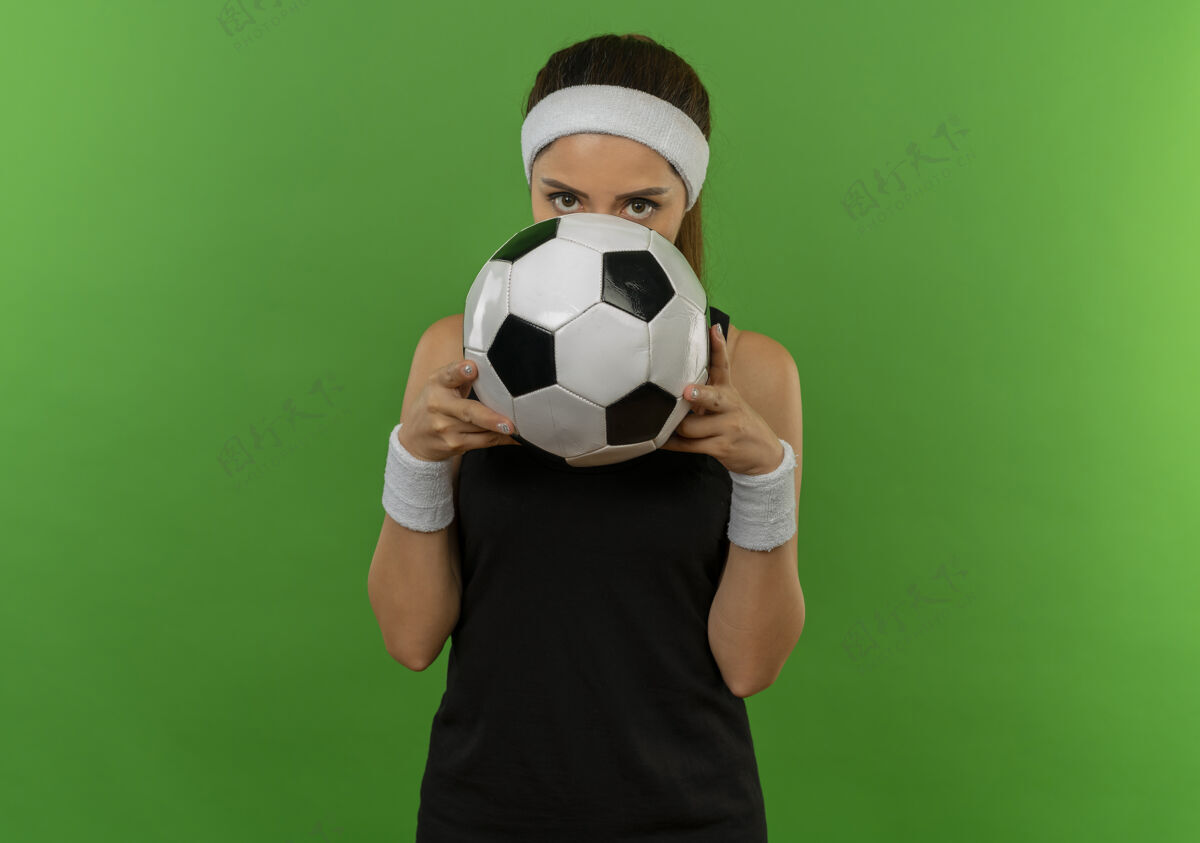 运动身穿运动服的年轻健身女 头戴足球带 把脸藏在足球后面 站在绿色的墙上偷看站运动员市民