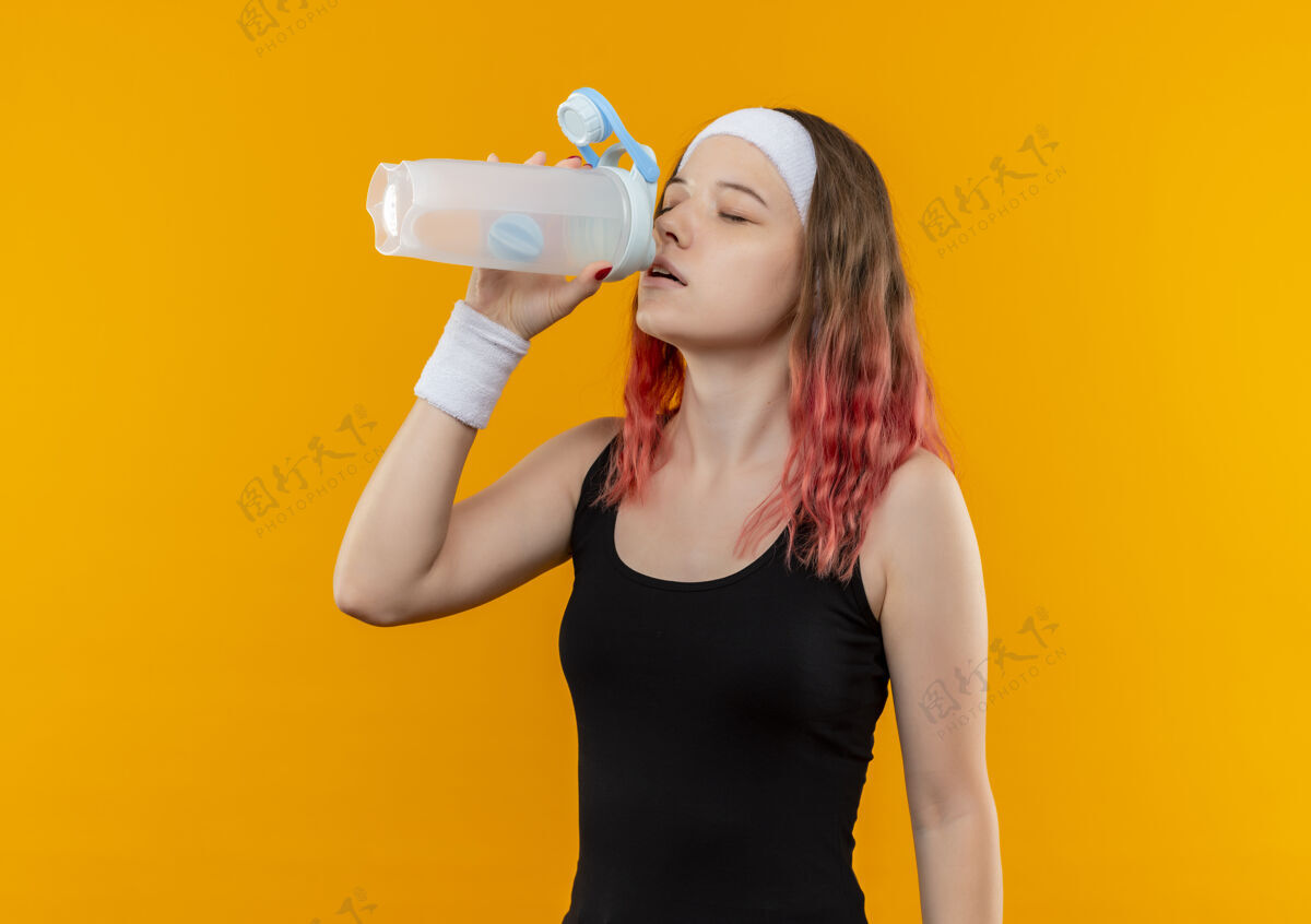 闭嘴穿着运动服的年轻健身女士闭着眼睛站在橙色的墙上喝水喝水运动装水