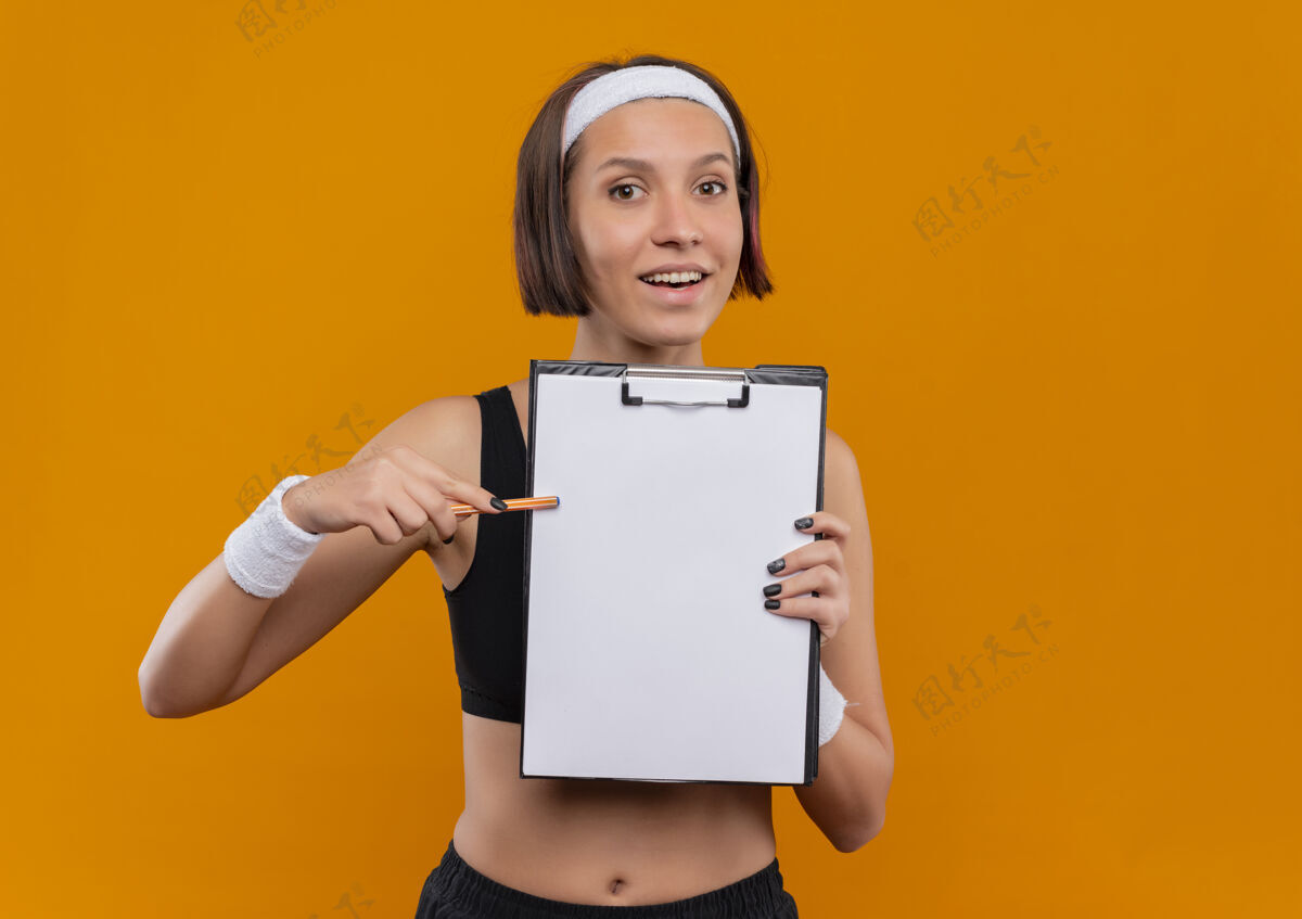 微笑身穿运动服的年轻健身女士 头上戴着头带 展示着空白页的剪贴板 用笔指着剪贴板 友好地微笑着站在橙色的墙上年轻人运动朋友