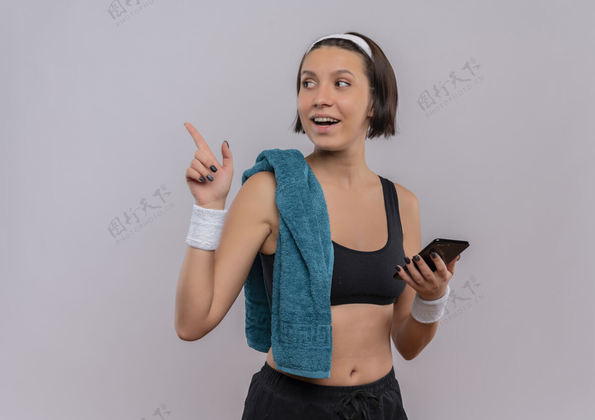 姿势身穿运动服的年轻健身女士 肩上挎着毛巾 手拿智能手机 手指指向一边 站在白色的墙上 开心地微笑着年轻人运动装毛巾