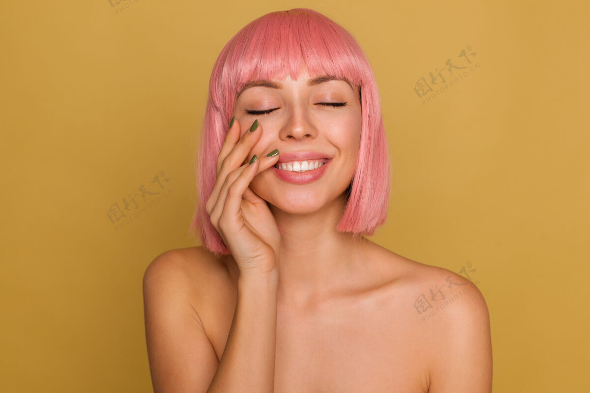 头发可爱漂亮的年轻粉红头发女性 自然妆容 举手轻抚脸庞 在芥末墙上摆姿势时 面带微笑肖像情感美甲