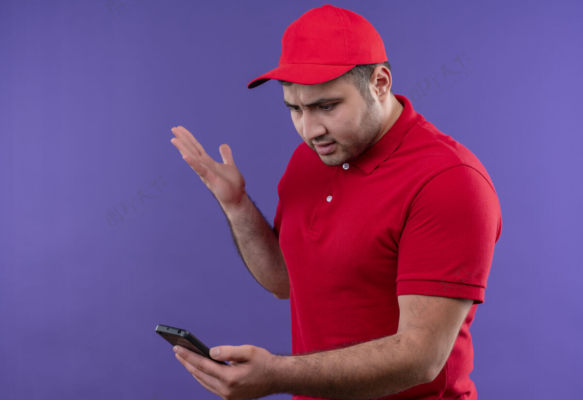 人年轻的送货员穿着红色制服 戴着帽子 站在紫色的路边 看着智能手机的屏幕 既不高兴又失望屏幕站不高兴
