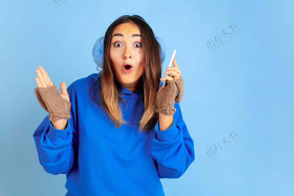 职业使用电话 快乐蓝色工作室背景上的白种女人肖像穿着暖和衣服的漂亮女模特人类情感的概念 面部表情 销售 广告冬天的心情 假期表情情绪手势
