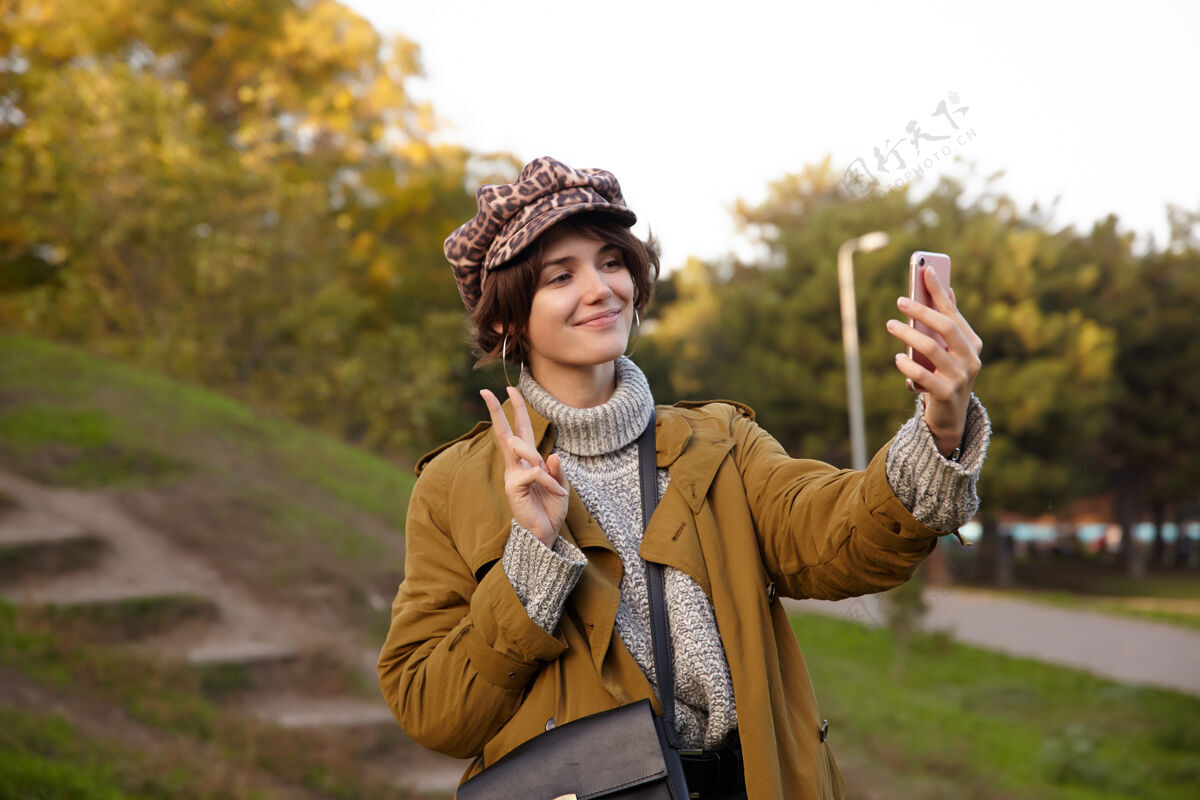 女性照片中 迷人的棕色头发的年轻女子鲍勃发型微笑着愉快地与她的手机自拍 举手与胜利的姿态 站在模糊的公园针织秋天20多岁