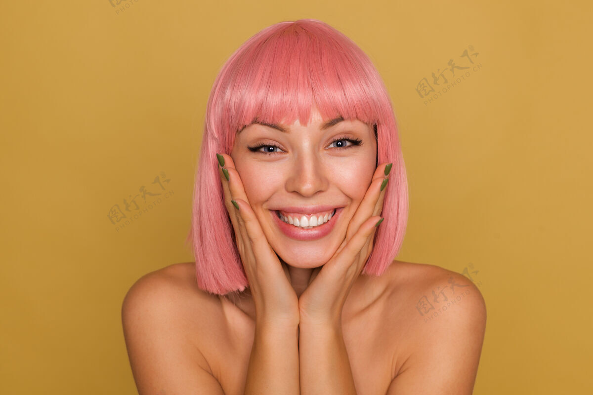 特写特写镜头：快乐的年轻蓝眼睛粉红色头发的女性 留着时髦的短发 手掌放在脸颊上 脸上洋溢着幸福的笑容 隔离在芥末墙上短裤情感休闲