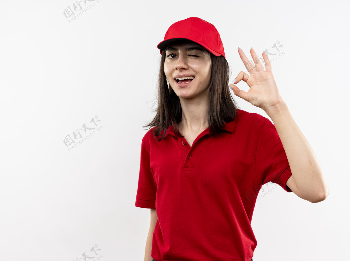 好身穿红色制服 头戴鸭舌帽的年轻送货员看着镜头 满脸笑容 在白色背景上闪烁着“ok”的手势站眨眼帽子