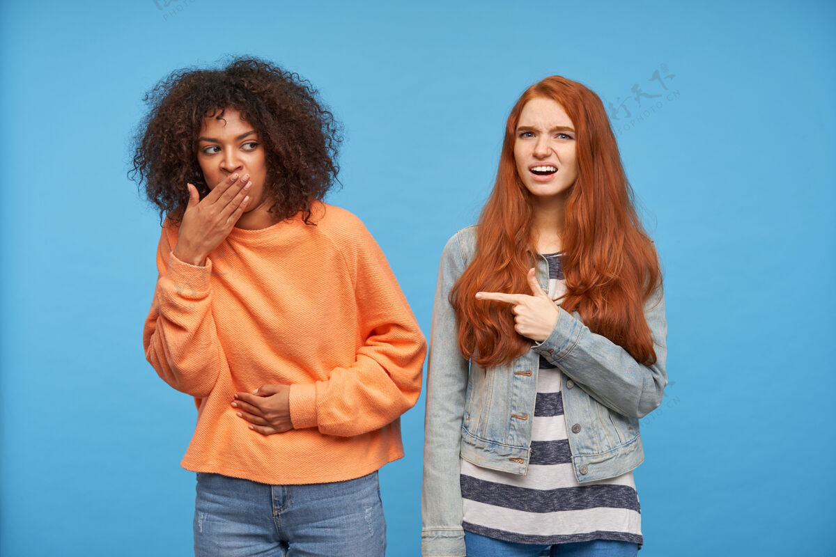 外套一张室内照片 年轻的棕色头发的黑皮肤女性 穿着橙色针织毛衣和牛仔裤 捂住嘴 手放在肚子上 和困惑的红发女士在蓝色的墙上合影女人红发尖