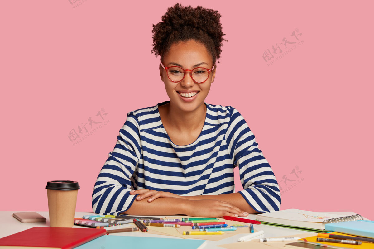 桌面一位快乐的黑皮肤女士 非洲裔头发梳成马尾辫 有着牙齿般的微笑 对工作的好成绩感到高兴 在记事本上创造了一幅图画 戴着红框眼镜 被隔离在粉色上计划学习日记