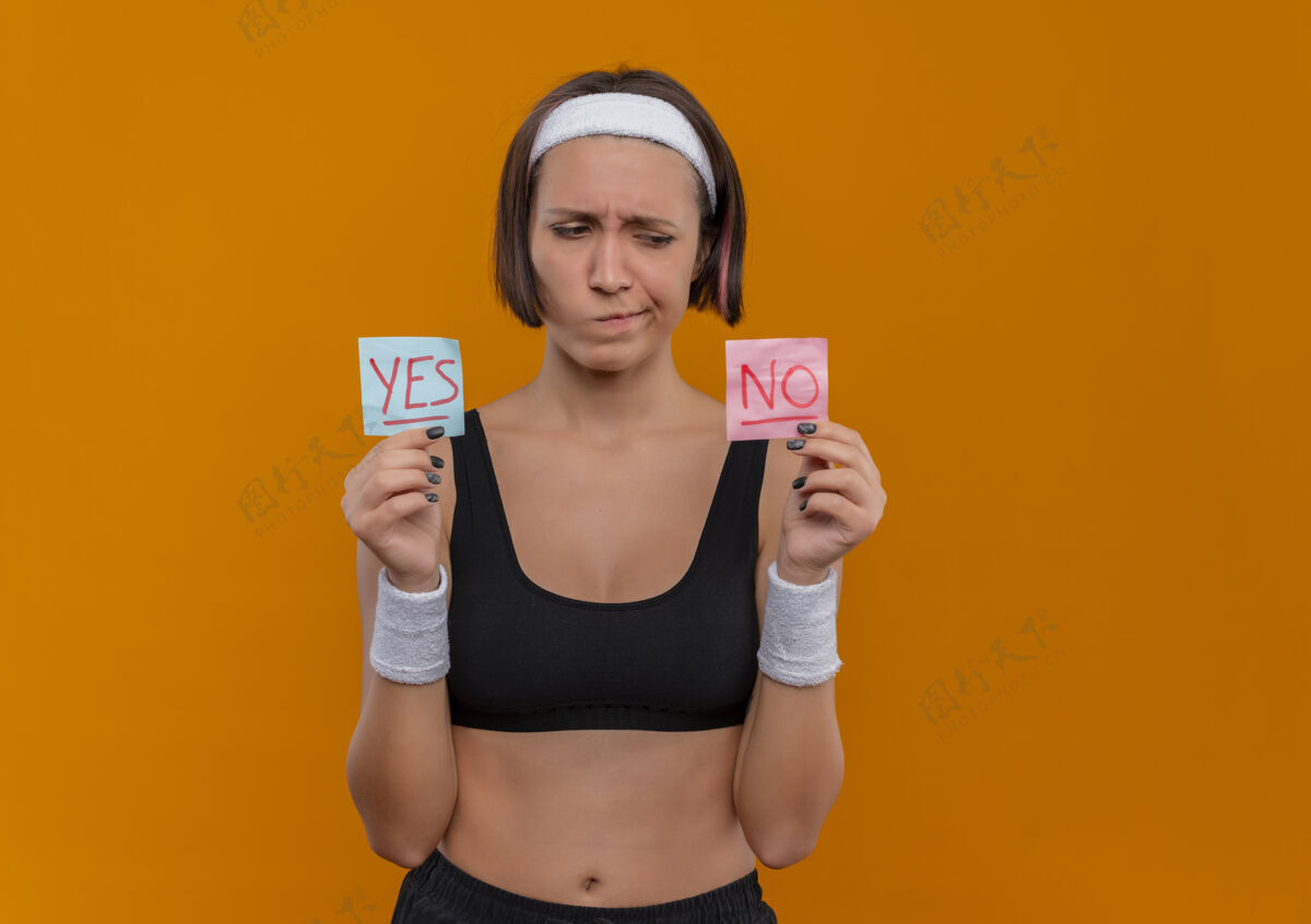 站着一位身穿运动服 戴着头巾的年轻健身女士举着两张写着“是”和“否”的提醒纸 站在橙色的墙上 带着怀疑的表情看着它们运动员女人运动
