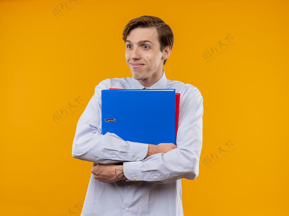 夹着身穿白衬衫的年轻人拿着文件夹 站在橙色背景下 面带腼腆的微笑向一旁望去衬衫脸害羞
