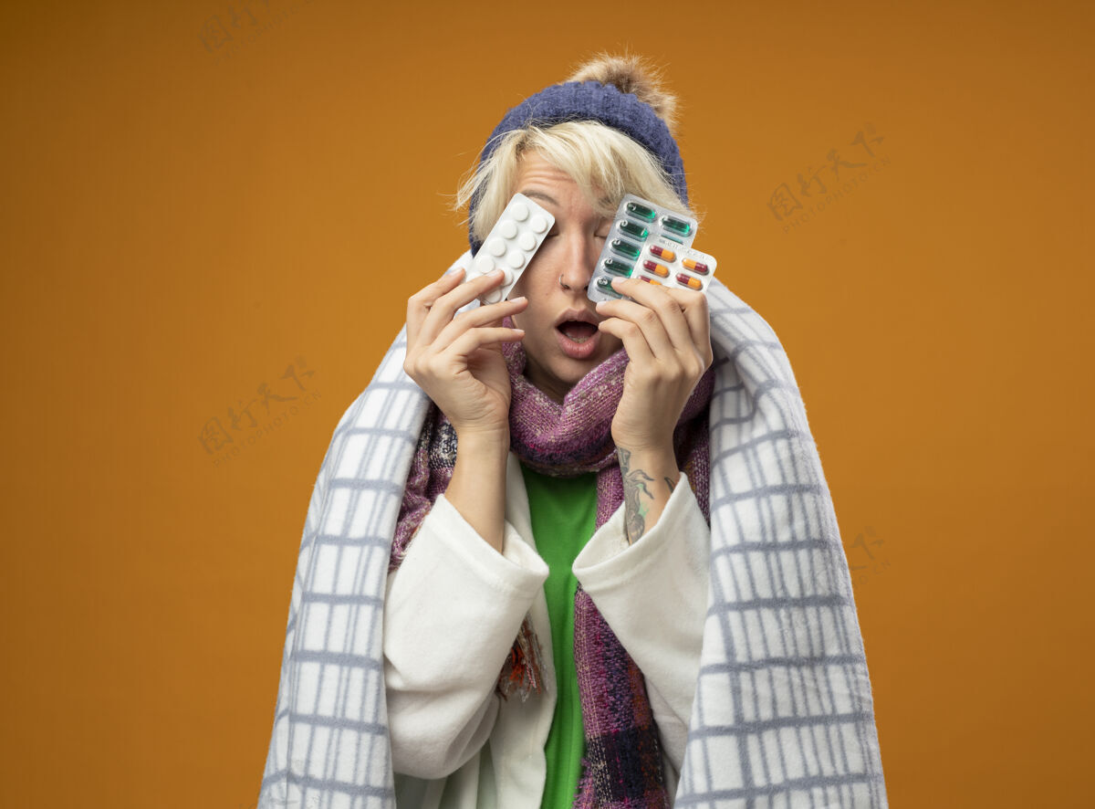 封面生病的不健康的女人 短头发 戴着暖和的围巾 帽子裹在毯子里 拿着包裹眼睛的带子 站在橙色的背景上包装毯子眼睛