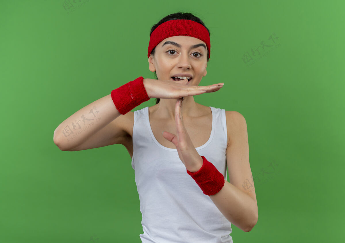 头带戴着头巾的年轻健身女 双手站在绿色的墙上 做着暂停的手势做运动手势
