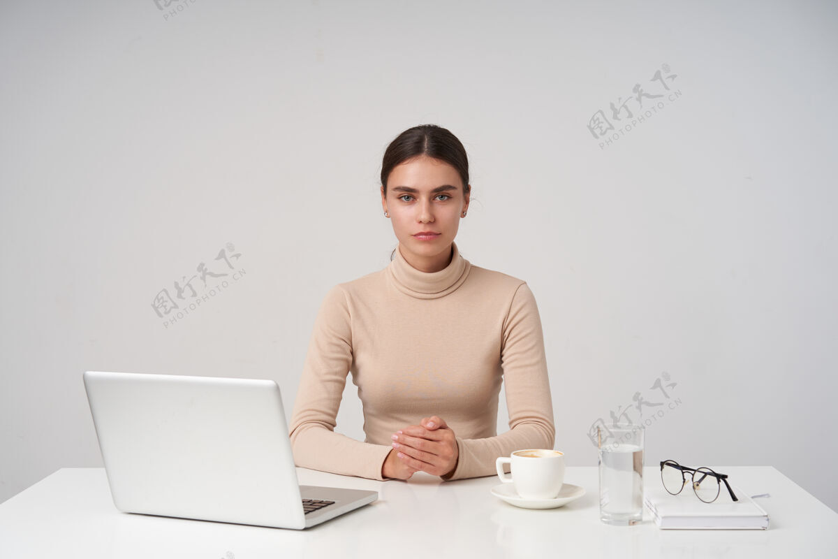 发型年轻迷人的黑发女商人坐在桌边 双手合十放在台面上 隔着白墙 穿着正装折叠工作肖像