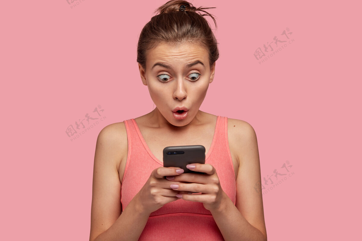 晒黑精神错乱的女人盯着智能手机 在网页上读新闻时感到震惊 连接到无线互联网 在健身房和教练训练后休息 穿着粉色上衣阅读肖像快递