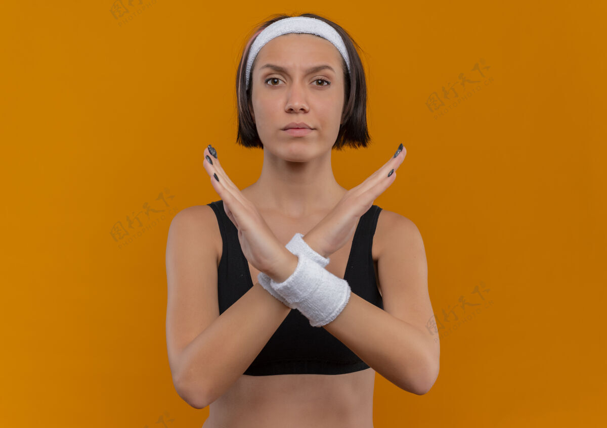 十字架身穿运动服 头箍交叉双手 站在橙色墙壁上做防御手势的年轻健身女士制造女人站立