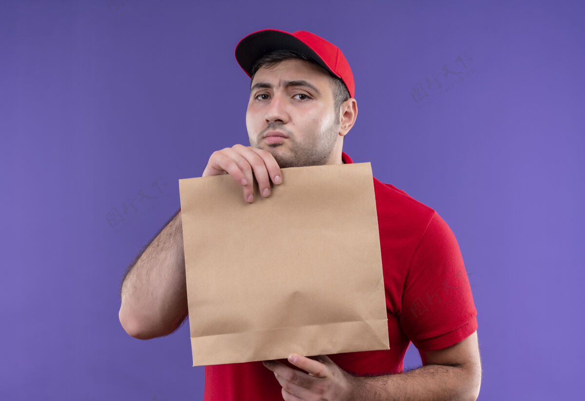 拿着胆小的年轻送货员穿着红色制服 戴着帽子 手里拿着一个纸包裹 站在紫色的墙上人人制服