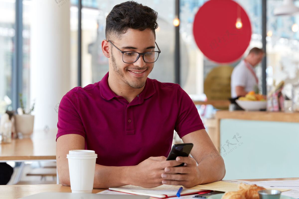青少年满意的自由职业者在线工作 持有手机 在社交网络中发送短信 坐在餐厅学生与groupmate交换通知 准备研讨会 写笔记企业家咖啡室内