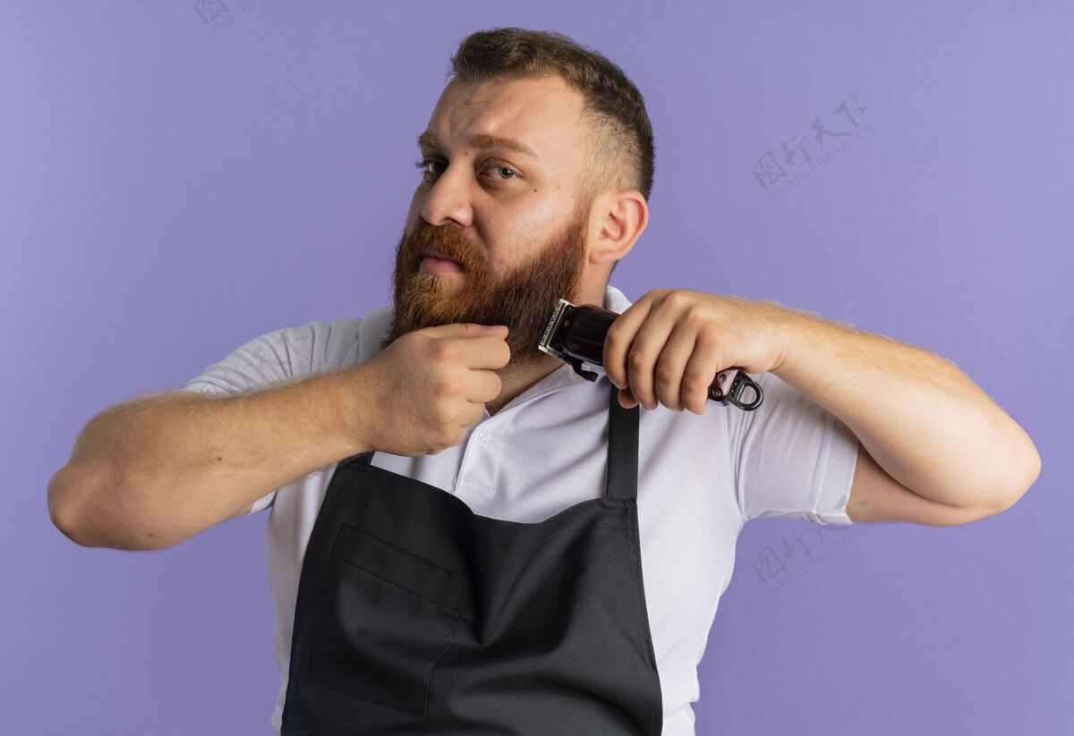 专业专业的胡须理发师站在紫色的墙上 围着围裙用剃须刀剪胡子 看上去很困惑困惑理发师男人