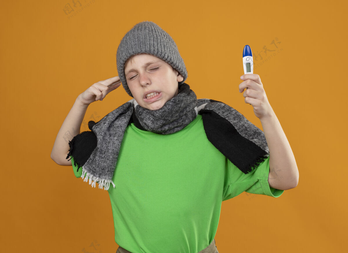 疾病生病的小男孩穿着绿色t恤 戴着暖和的围巾 戴着帽子 显示体温计 感觉不舒服 生病 不开心 站在橙色背景下 在寺庙附近做手枪手势手势帽子男孩