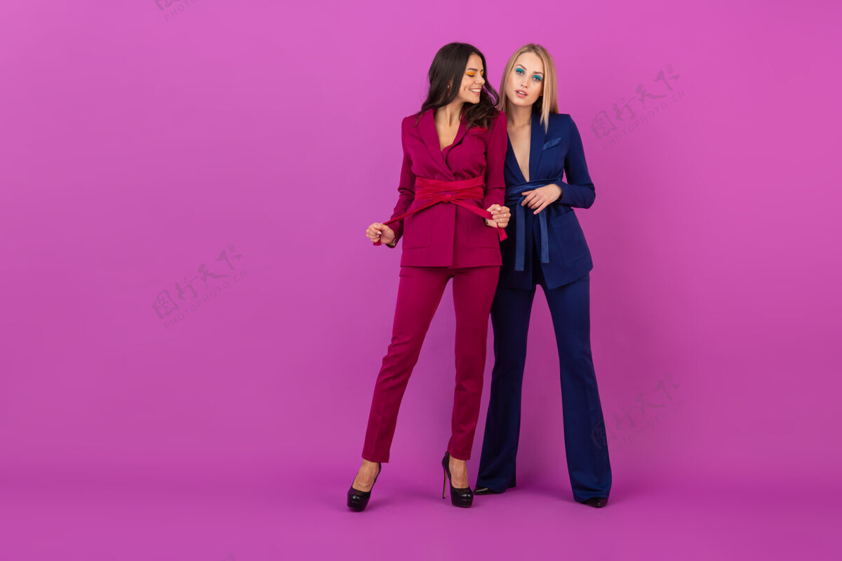 秋天高级时尚风格两位微笑迷人的女士在紫罗兰色的墙壁上穿着时尚多彩的紫色和蓝色晚礼服 朋友们一起玩 时尚潮流休闲华丽时尚