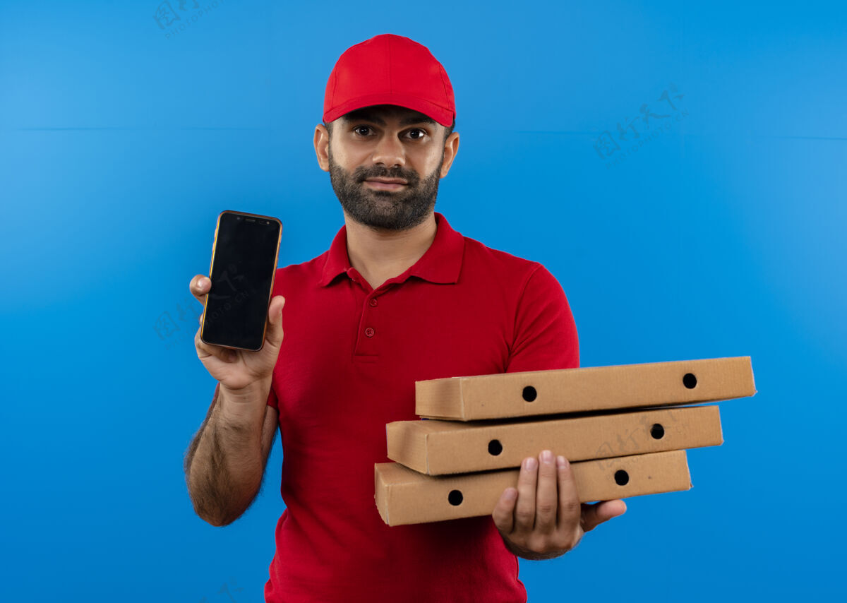 自信有胡子的送货员穿着红色制服 戴着帽子 手里拿着一叠披萨盒 站在蓝色的墙上 脸上露出自信的微笑叠着站着送货