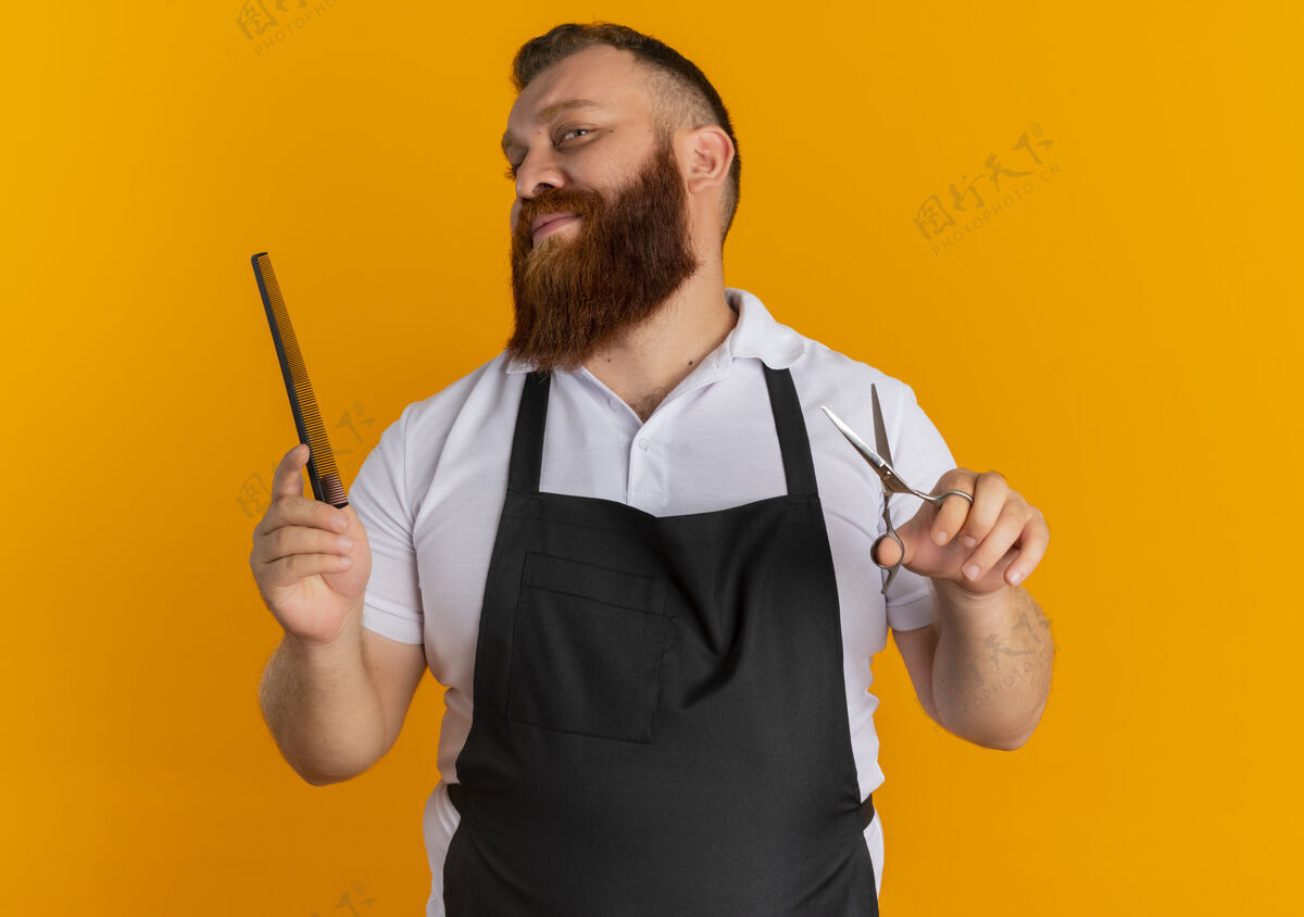 男人专业的胡须理发师站在橙色的墙上 围着围裙 手里拿着剪刀和发刷 脸上带着微笑胡子脸微笑