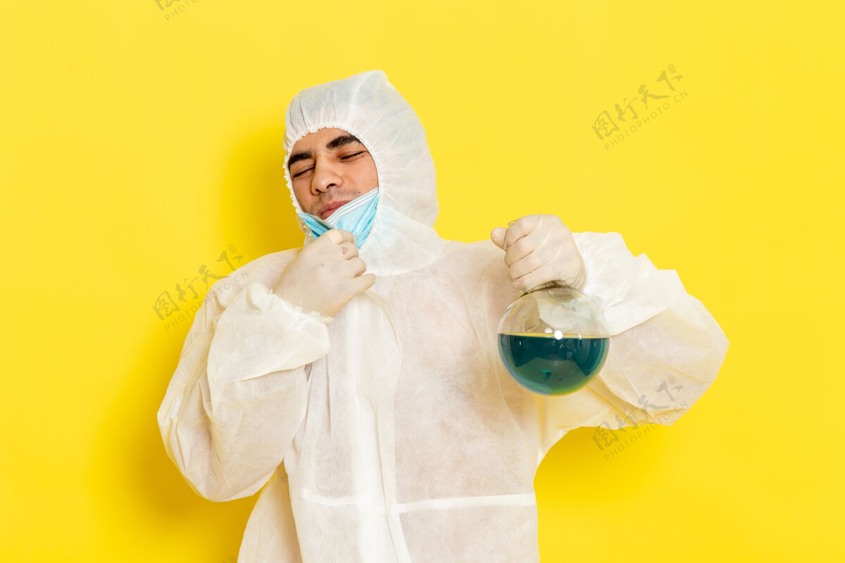 化学正面图穿着特殊防护服的男科学工作者拿着装有蓝色溶液的烧瓶 在黄色的表面上摘下面具服装男性手持