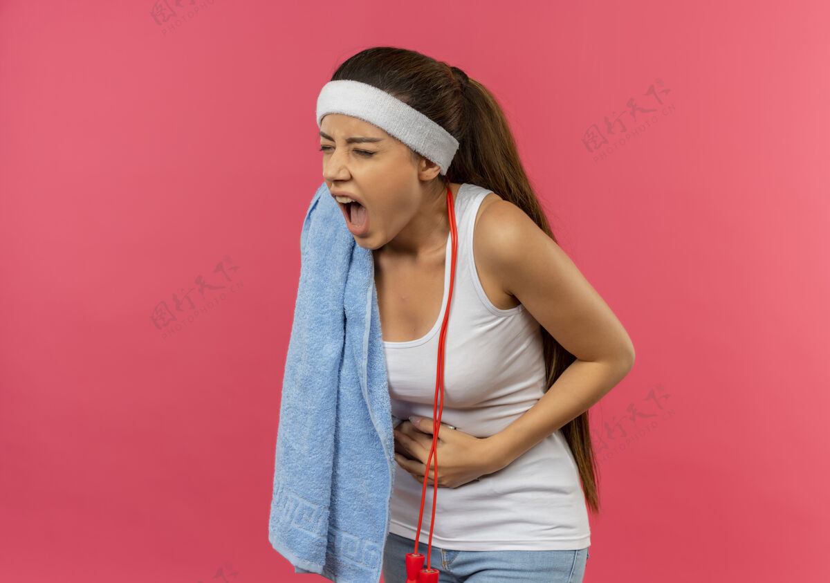 看穿着运动服的年轻健身女 头上戴着头巾 肩上搭着毛巾 摸着肚子 站在粉色的墙上 看上去不舒服 感觉疼痛疼痛毛巾头带