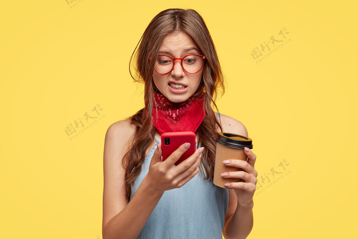 负片不满意的可爱女人在手机上厌恶地看着 在专门的应用程序里编辑照片 喝咖啡去 在黄色的墙上摆姿势 感到厌恶 连接到高速互联网短信眼镜白种人