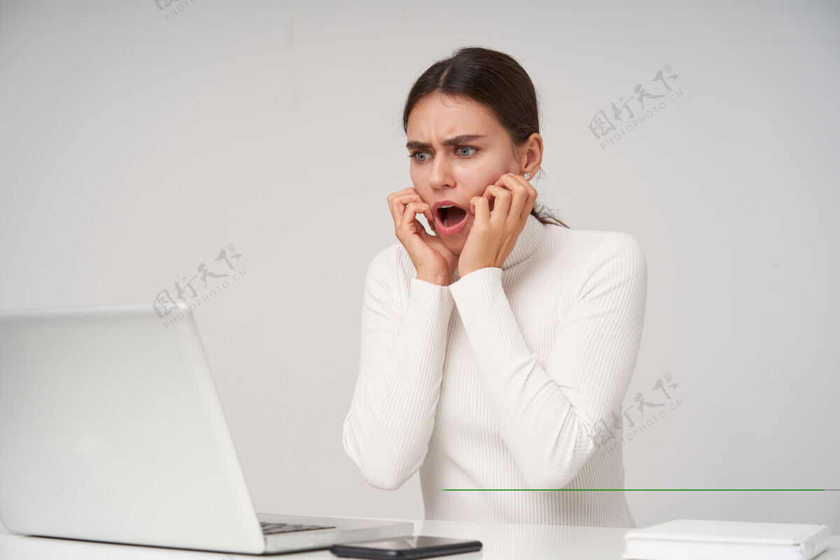 工作震惊的年轻漂亮的黑发女人 自然妆容保持举起双手放在脸上 同时惊恐地看着她的笔记本电脑屏幕 隔着白墙漂亮女人欧式