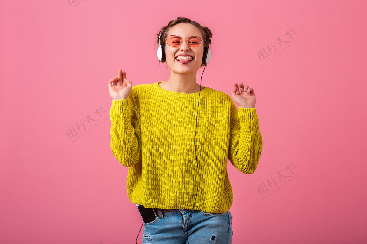享受快乐搞笑的女人戴着耳机听音乐穿着时髦的五颜六色的服装隔离在粉色的墙上 穿着黄色的毛衣和太阳镜 尽情展示舌头明亮声音有趣