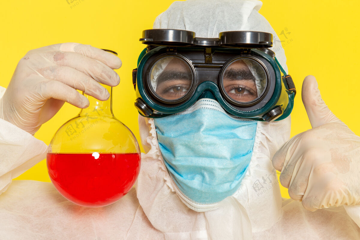 男性正面近距离观察穿着特殊防护服 戴着面罩的男性科学工作者 黄色桌子上放着装有红色溶液的瓶子服装科学化学