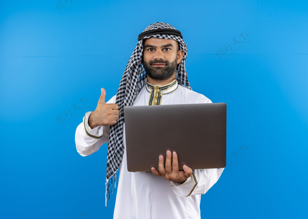 拿着身着传统服装的阿拉伯商人手持笔记本电脑微笑着竖起大拇指站在蓝色的墙上微笑显示穿